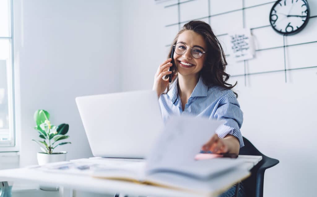 Laughing female multitasking speaking on smartphone and working with notepad in office
